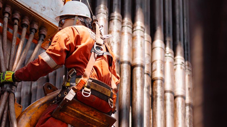 Oil field worker in orange overalls suspended on some pipework.