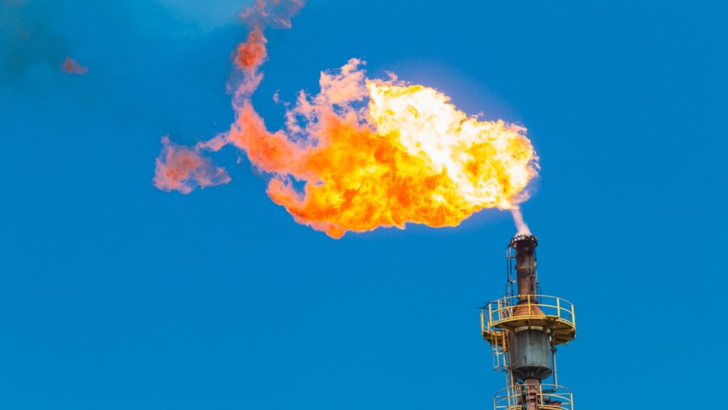 A flame coming from the top of a chimney on an oil field with a blue sky behind it.
