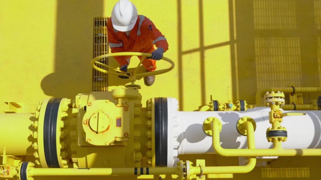 An engineer working on a pipeline of an oil field. The floor and some of the pipe are vivid yellow, contrasting against the orange of the worker's high vis protective clothing and white hard hat.