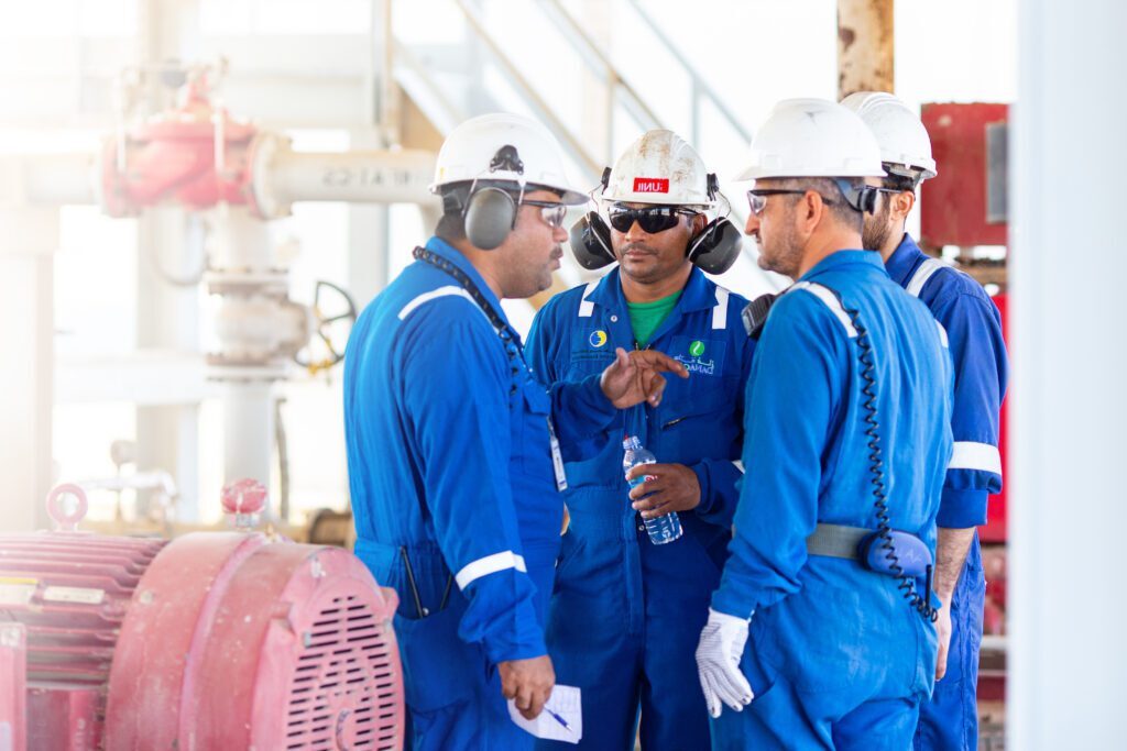 Four male engineers in blue overalls and white hard hats gather to discuss their work on an oil field.