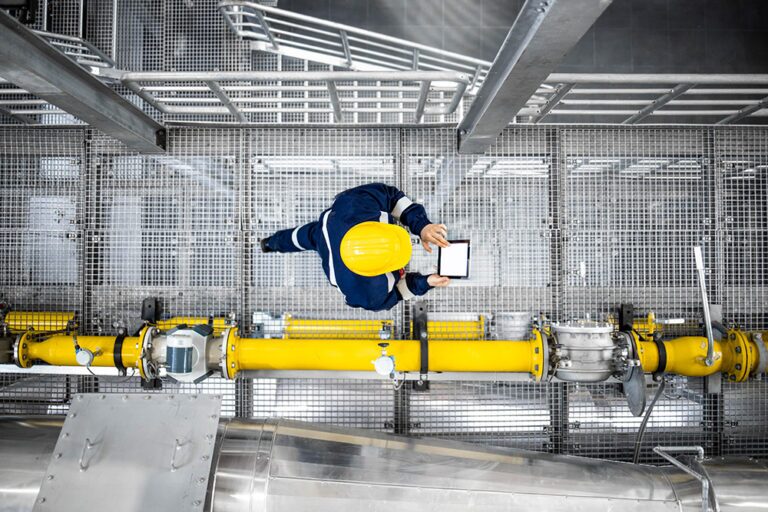 An areal shot of an engineer in dark overalls and a yellow hard hat walking through an oil facility whilst working on a hand held device. There is a bright yellow pipe running alongside them.