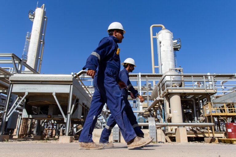 Two engineers in two blue overalls wearing white helmets walking in an oil & gas plant