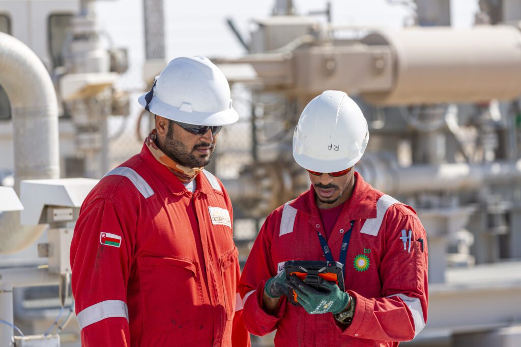 Two male engineers wearing red BP overalls and white hard hats working on a hand held device at a gas field in Oman.
