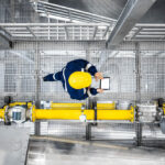 An areal shot of an engineer in dark overalls and a yellow hard hat walking through an oil facility whilst working on a hand held device. There is a bright yellow pipe running alongside them.