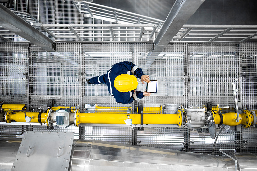 An areal shot of an engineer in dark overalls and a yellow hard hat walking through an oil facility whilst working on a hand held device. There is a bright yellow pipe running alongside them.